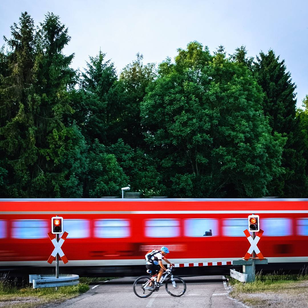 A cyclist passes a railway crossing as a red train blurs across a background of green trees.