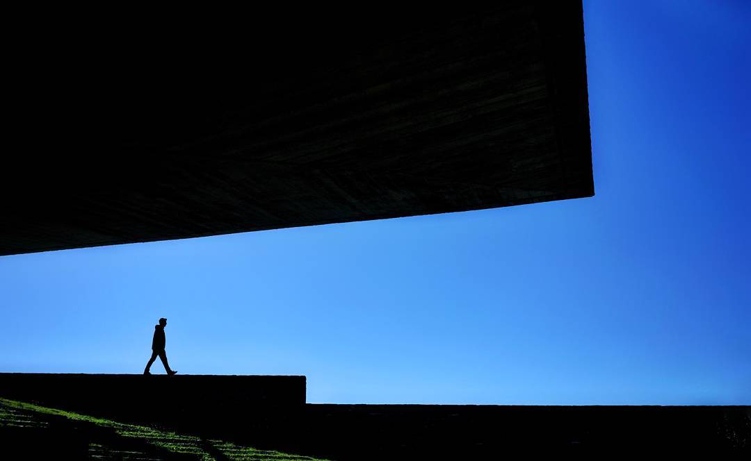 A small walking silhouette beneath a large angular roof against a vivid blue sky.